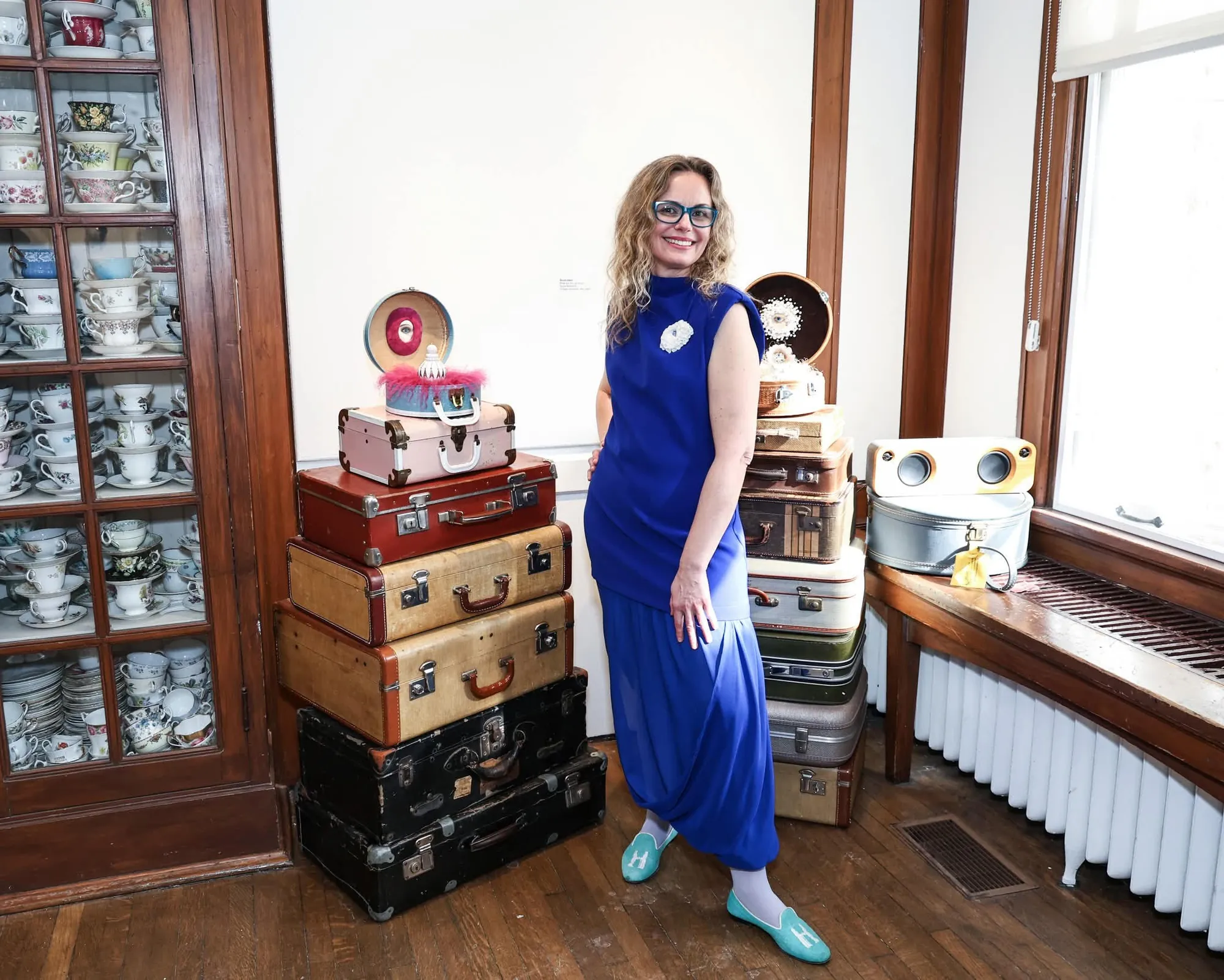 Tanya Besedina in a blue dress standing beside stacked vintage suitcases with ceramic sculptures on top, a china cabinet with teacups visible on the left