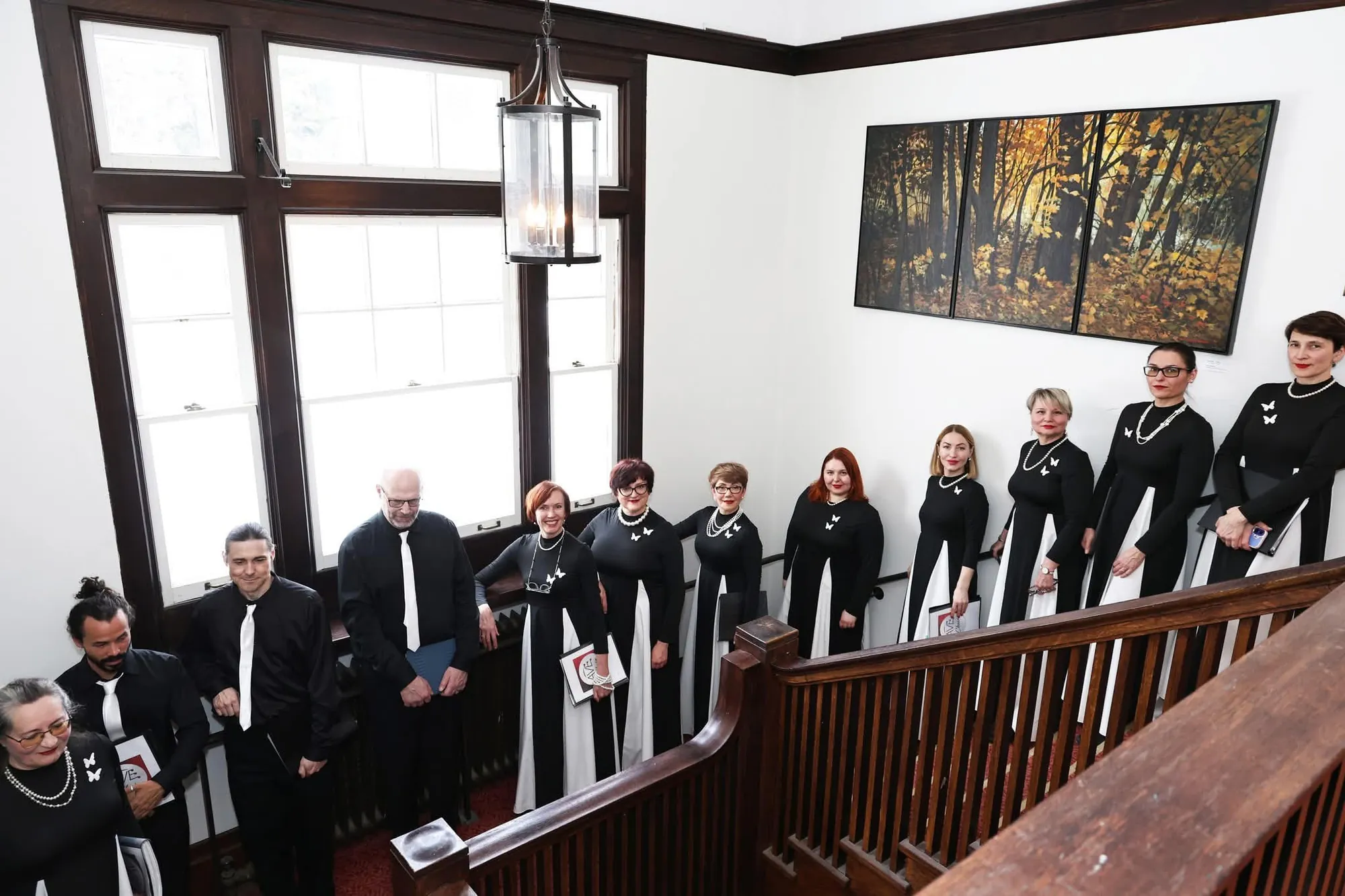 AVE Choir members posed on a staircase landing, photographed from above, wearing black outfits with white butterfly pins