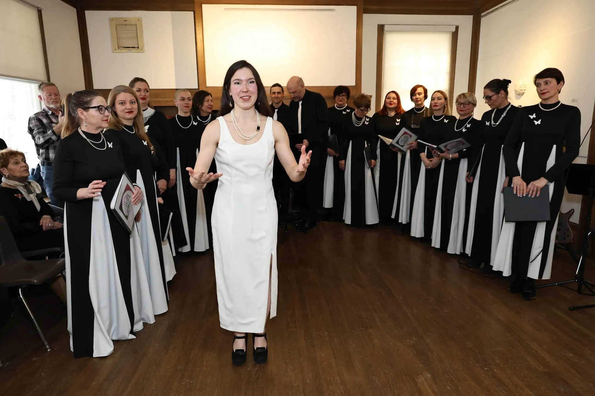 Wide shot of the full AVE Choir in a semicircle with the soloist in white at the center, audience visible to the side
