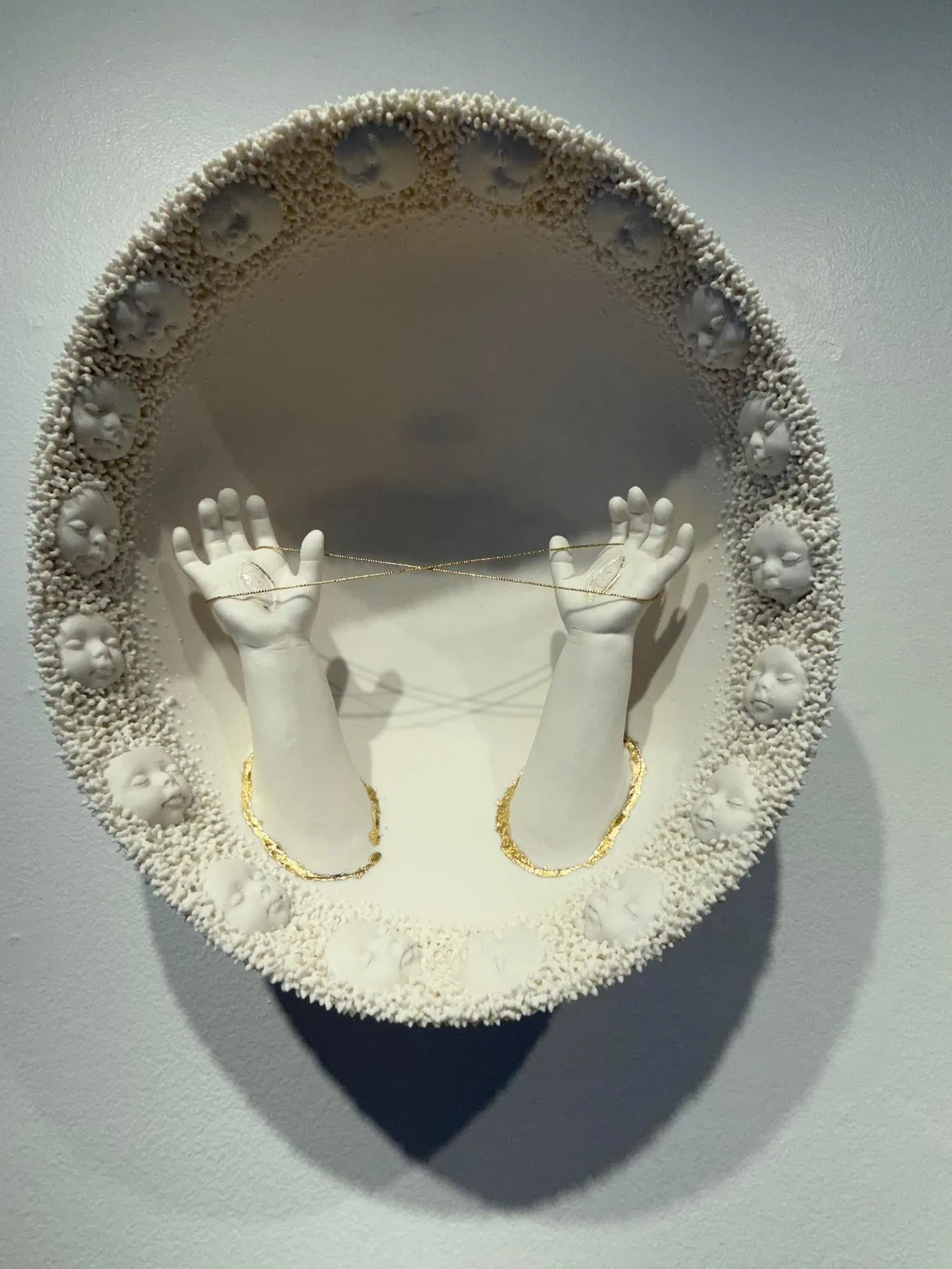 Oval porcelain wall sculpture with a textured border of child faces and two raised hands connected by a delicate gold chain