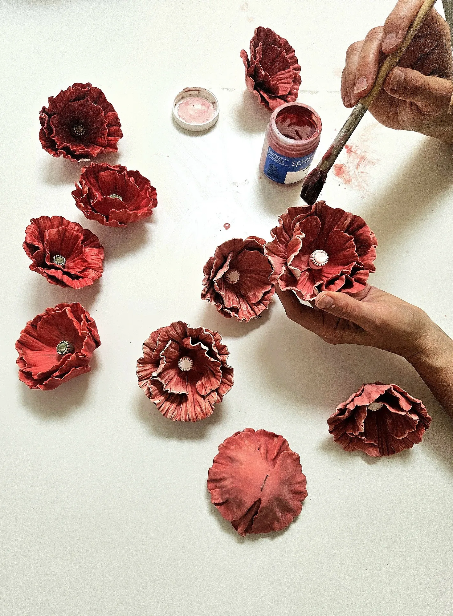 Hands painting multiple red ceramic poppies with a brush, a paint jar visible beside them