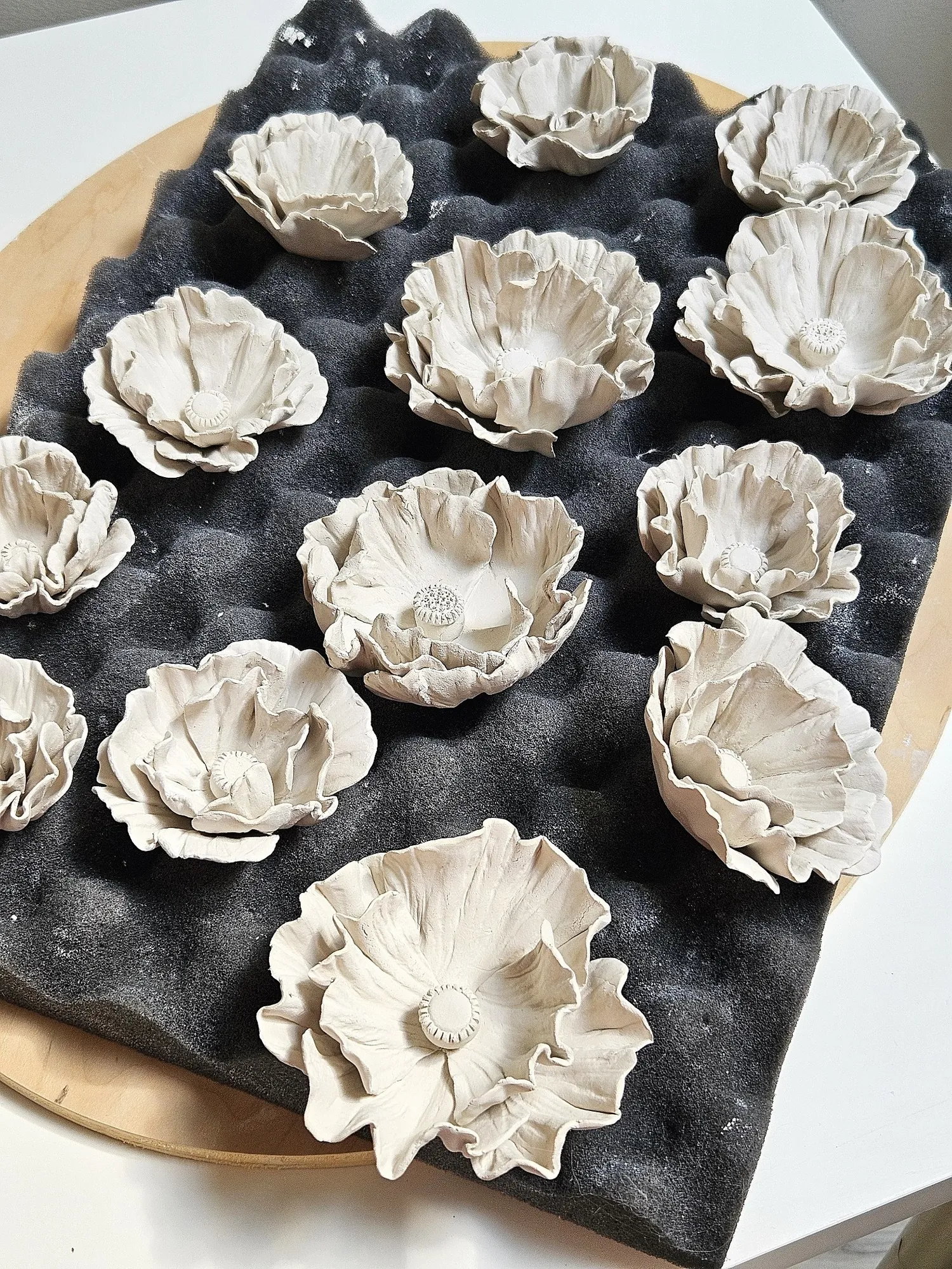 Many unfired white ceramic poppies drying on dark foam padding on a wooden board