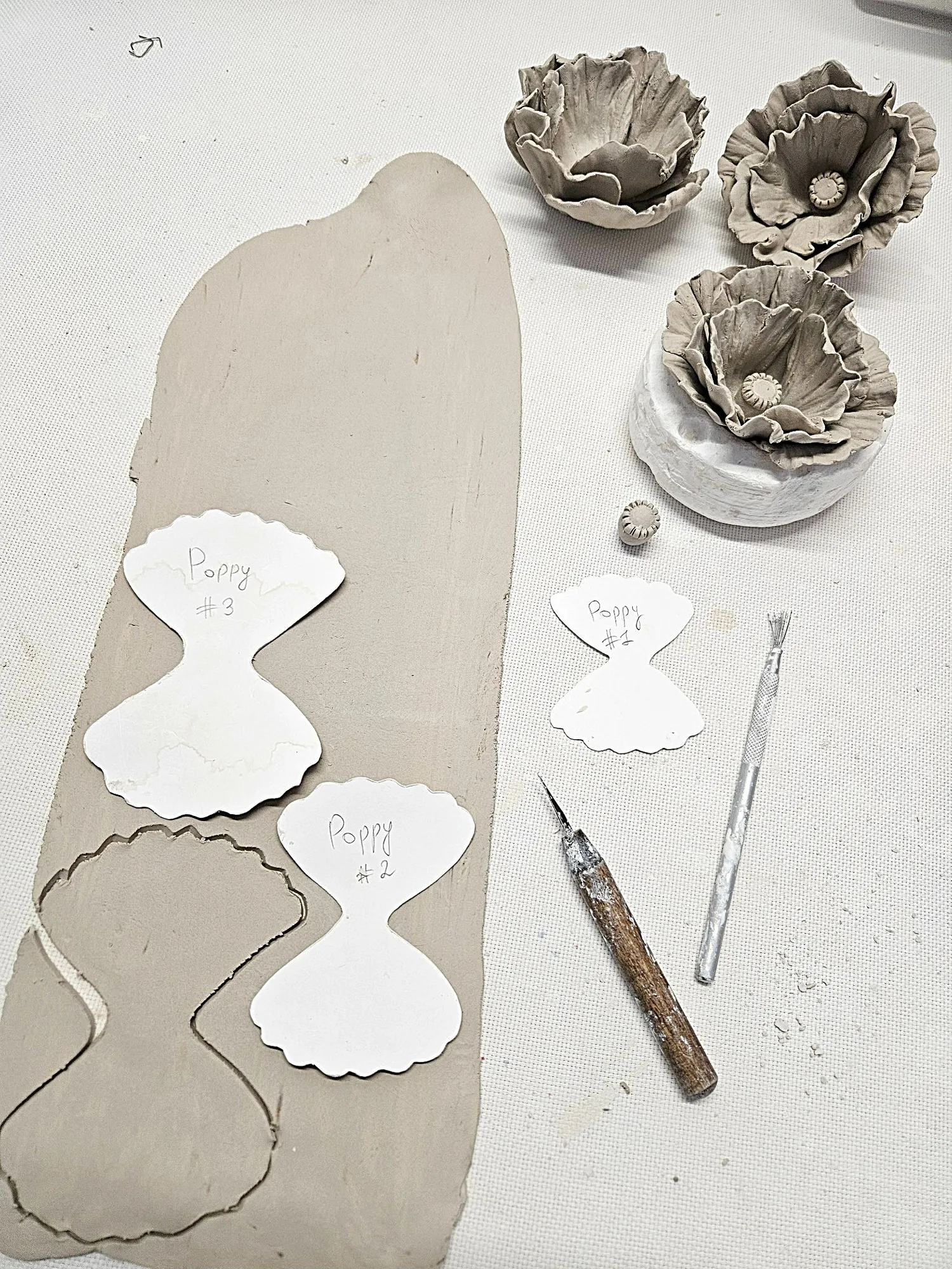 Clay slab with paper petal templates labeled Poppy 1, 2, and 3, alongside sculpting tools and unfired clay flowers